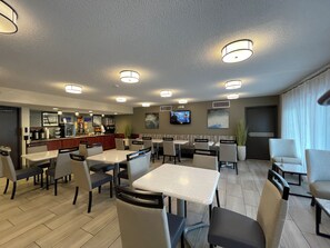 A hotel restaurant with multiple tables and chairs, a bar area, and a television screen on the wall.