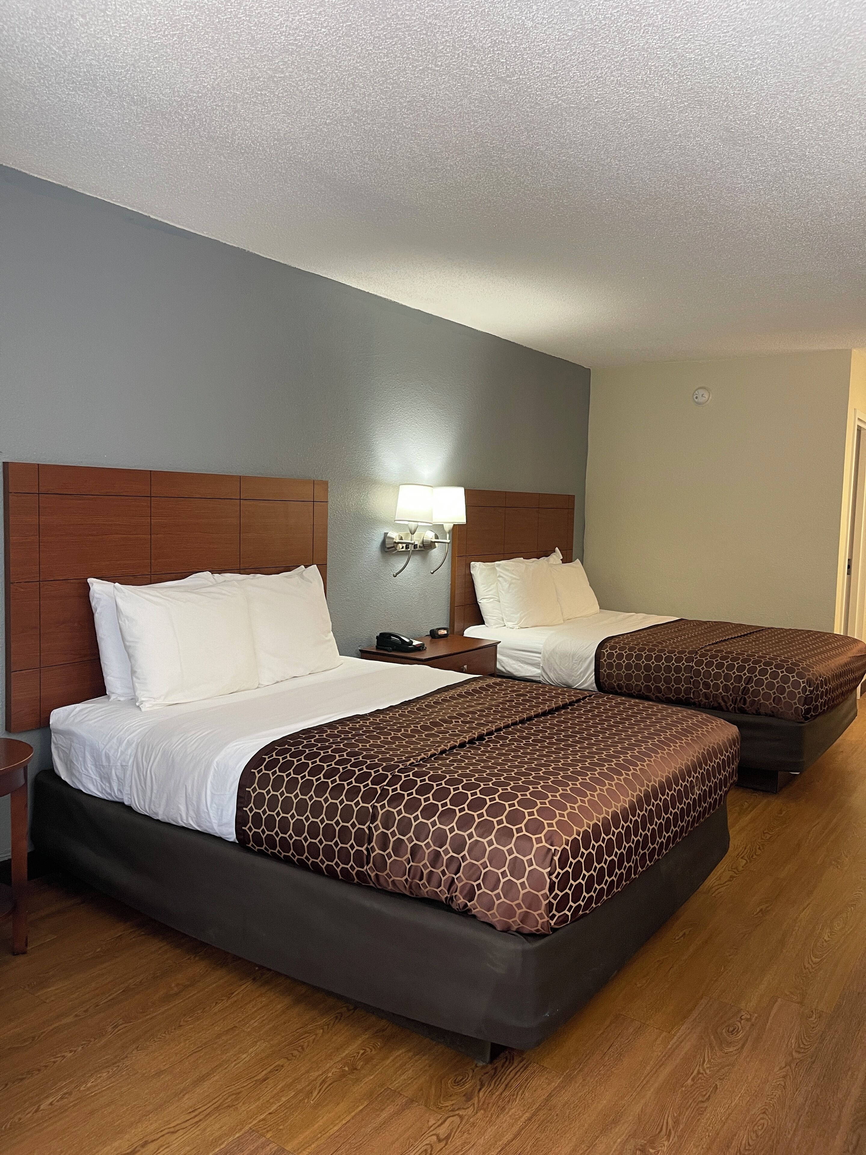 A hotel room with two beds, wooden headboards, a nightstand with a lamp, and a wooden floor.
