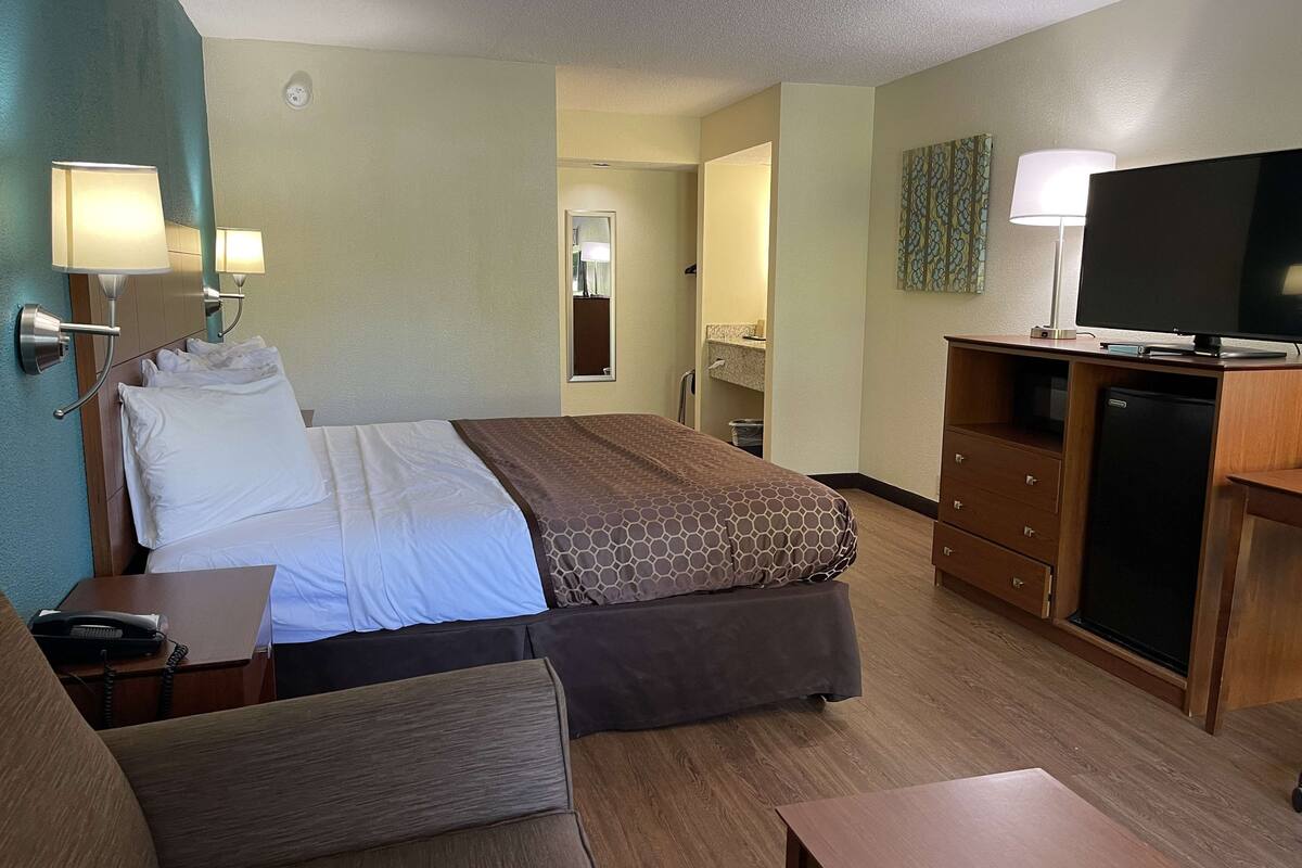 Ein Hotelzimmer mit Bett, Nachttisch, Lampe, Fernseher und Schreibtisch mit Computer.