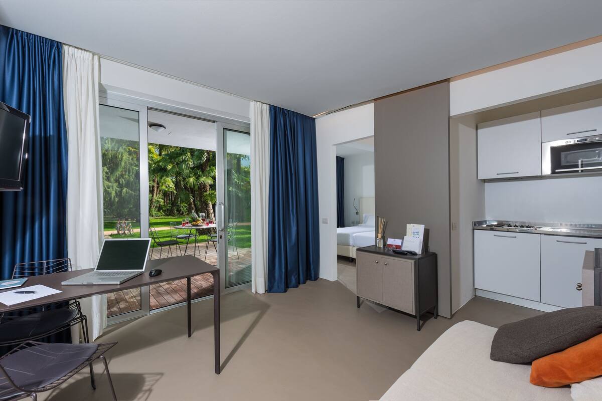 A hotel room with a desk, laptop, and a TV. There is a balcony with curtains and a view of greenery. The room has a kitchenette with a microwave and a stove.