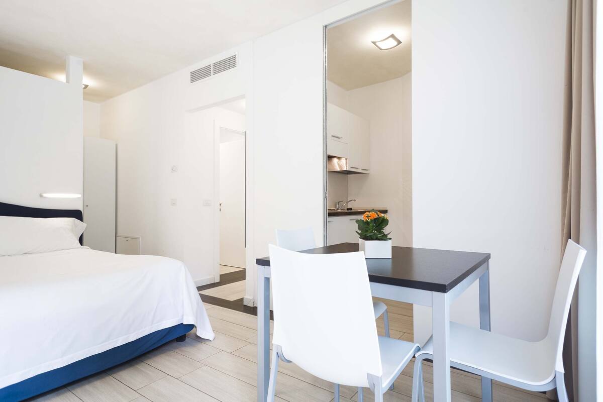A hotel room with a bed, a desk, two chairs, a small table with a flower, and a kitchen area in the background.