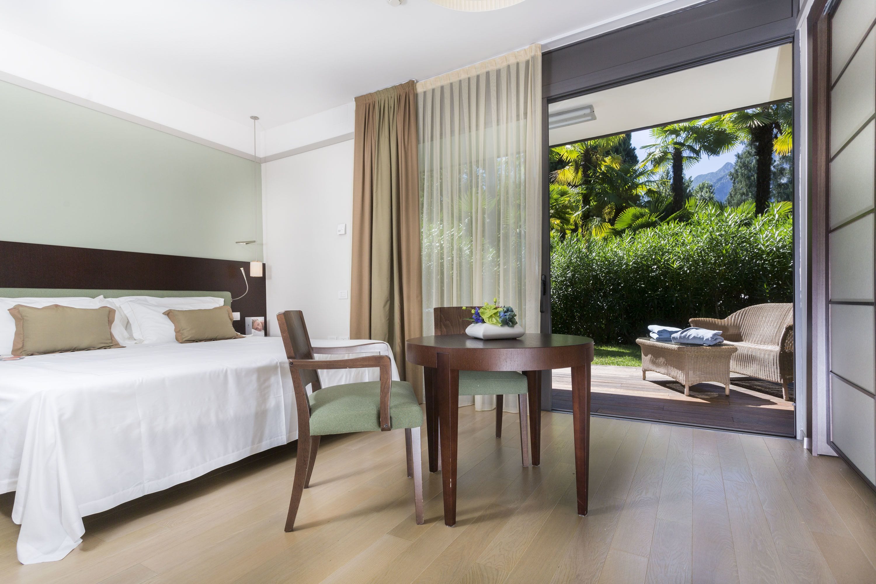 Ein Hotelzimmer mit Bett, Sessel, Tisch und Blick auf einen Garten durch eine Schiebetür.