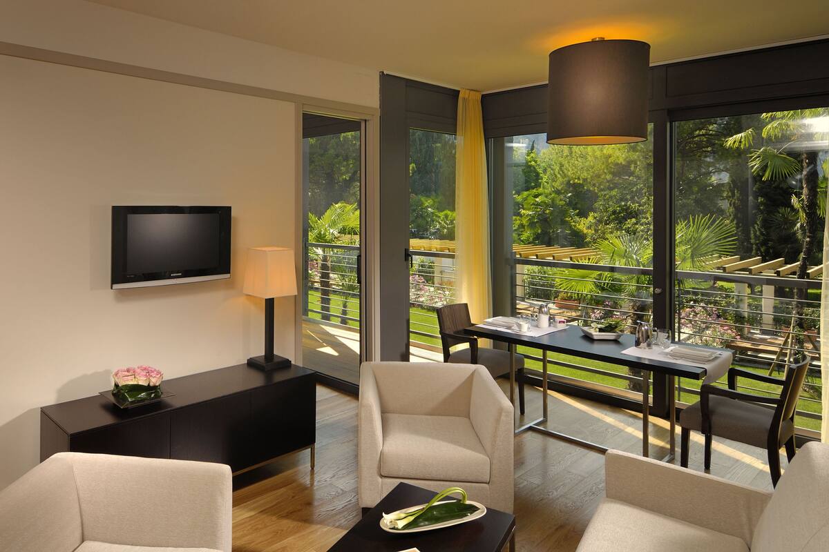A modern hotel room with a flat-screen TV, a bedside table with a lamp, a sofa, and a dining area with a table and chairs.