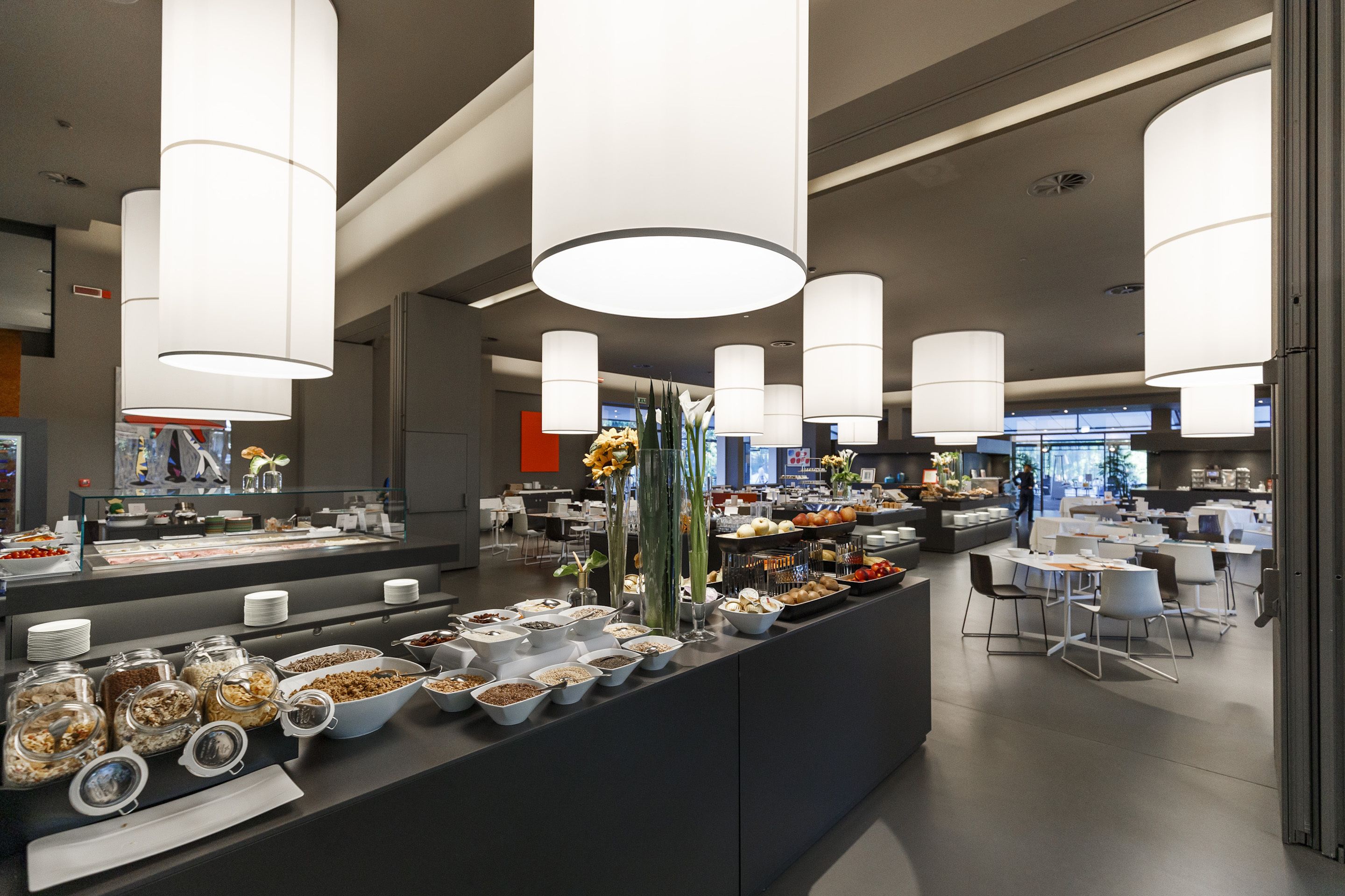 Un espace buffet moderne doté de plusieurs points de service, proposant une variété de plats et de boissons, ainsi qu’un espace repas avec des tables et des chaises.