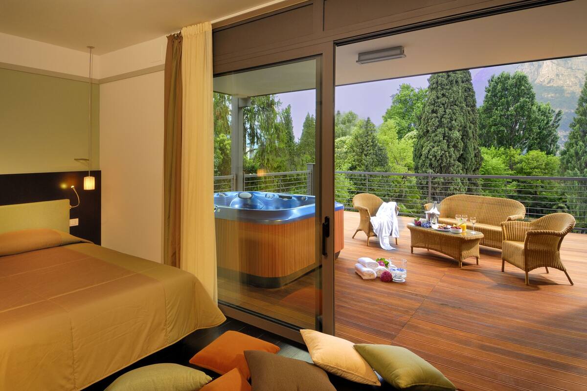 A hotel room with a bed, a hot tub, and a balcony with wicker furniture and a view of trees.