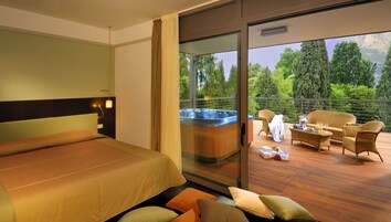 Habitación de hotel con cama, bañera de hidromasaje y balcón con mobiliario de mimbre y vistas a árboles.