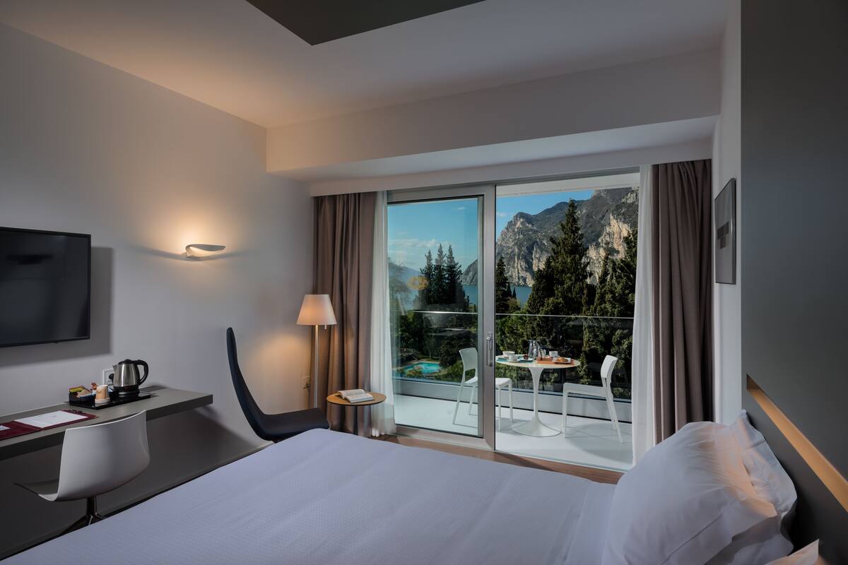 A modern hotel room with a large bed, a desk with a chair, a TV, a lamp, and a balcony with a pool and mountain view.