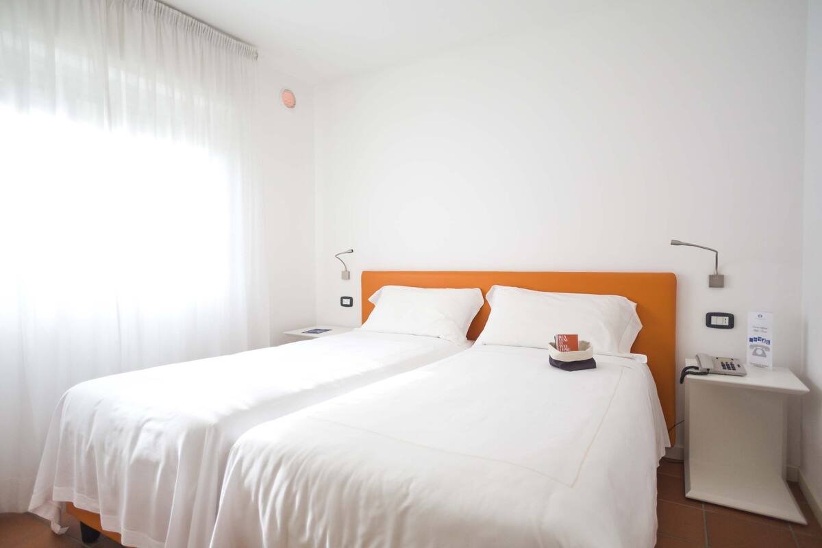 A hotel room with two single beds, white bedding, an orange headboard, a bedside table with a phone, and a window with sheer curtains.