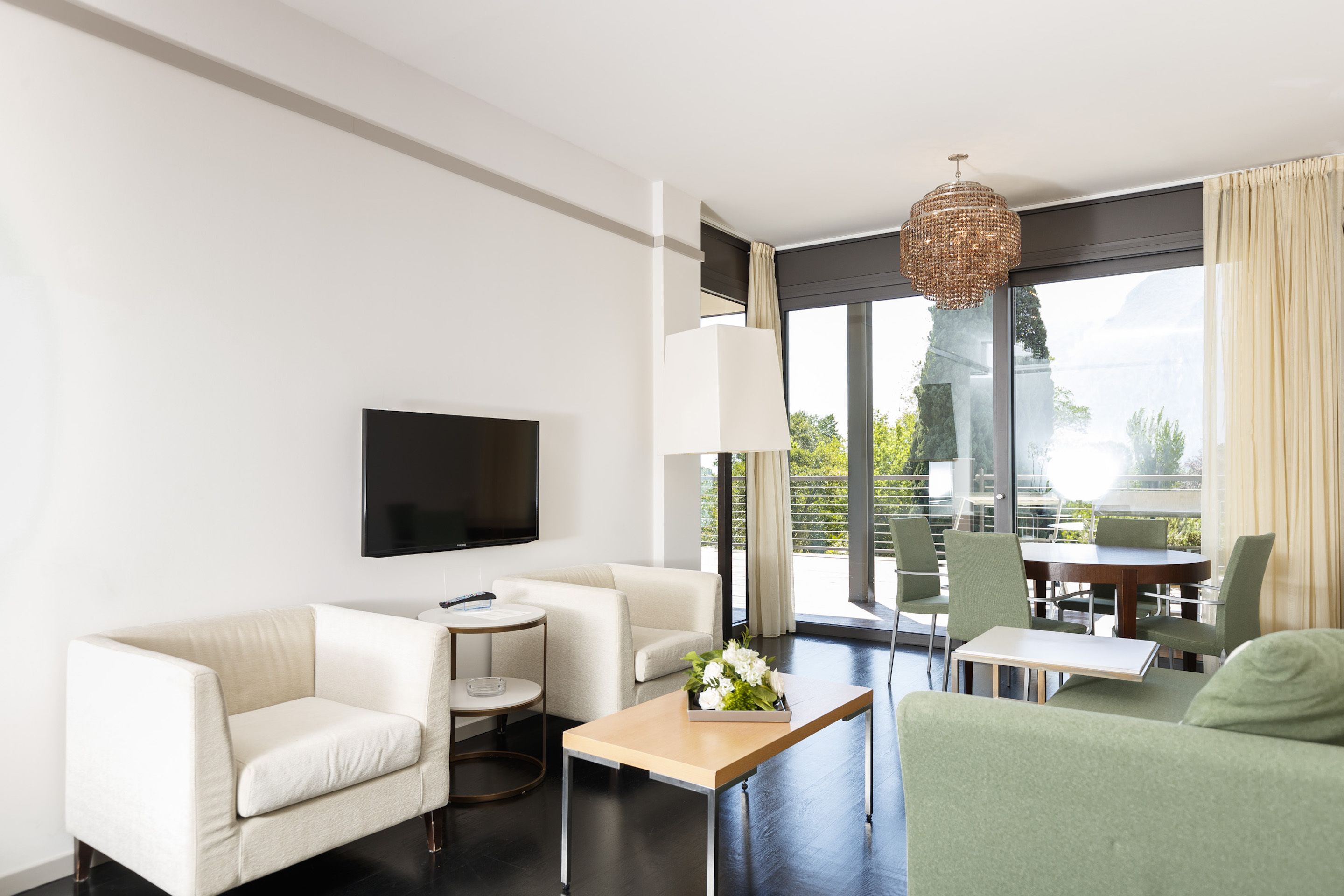 A modern hotel room with a flat-screen TV, a sofa, a small round table, a coffee table, and a balcony with a dining area.