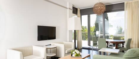A modern hotel room with a flat-screen TV, a sofa, a small round table, a coffee table, and a balcony with a dining area.