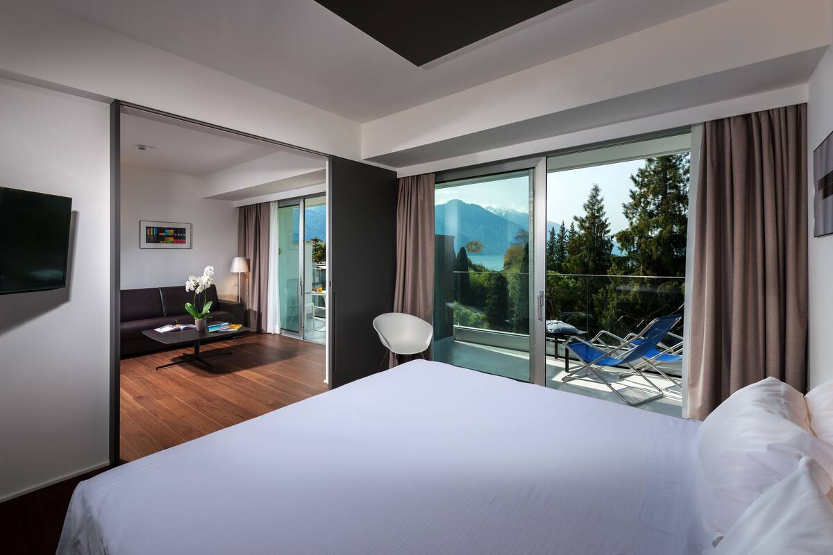 A modern hotel room with a large bed, a flat-screen TV, a sofa, a dining area with a table and chairs, and a balcony with a view of mountains and trees.