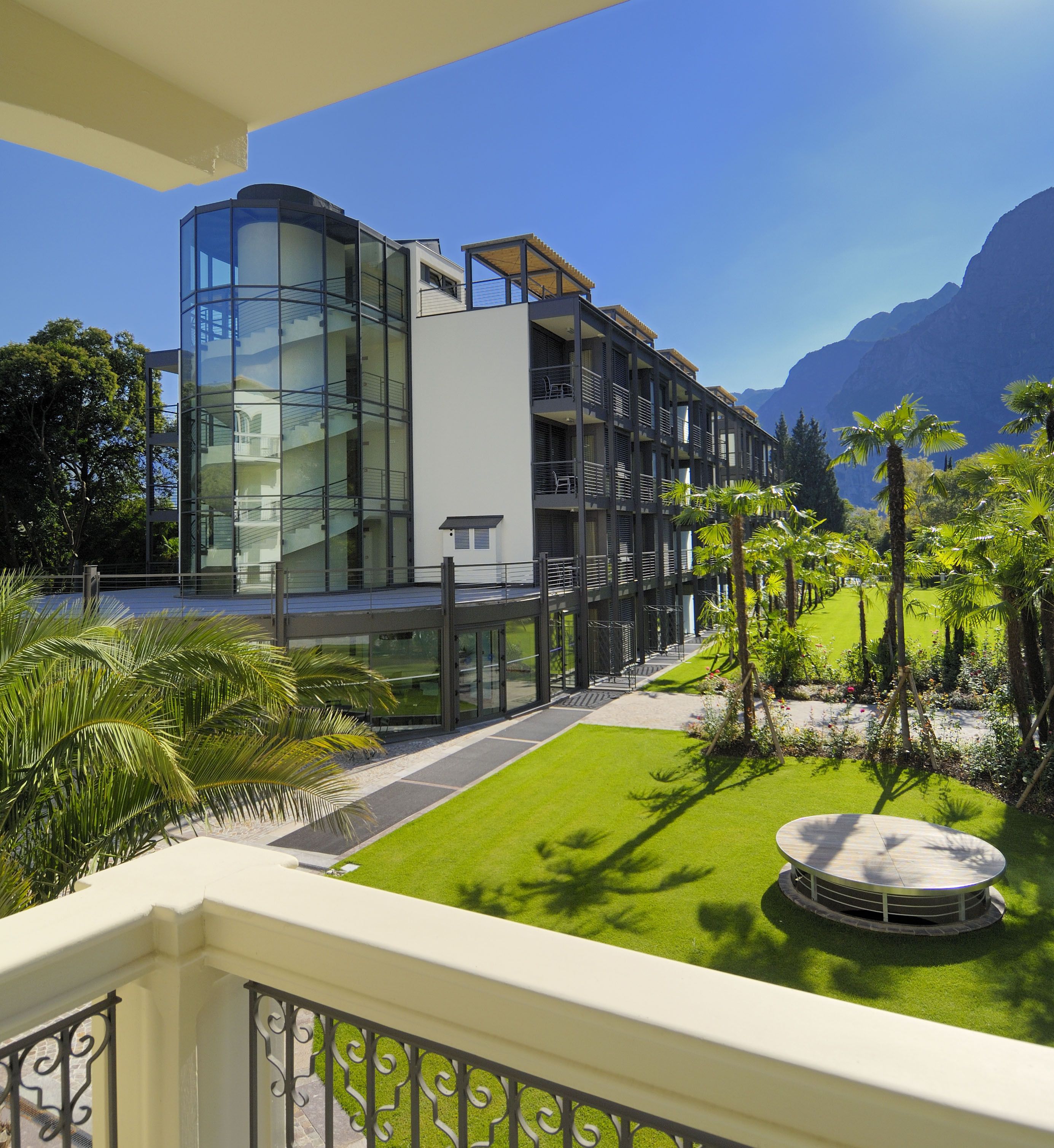 A modern hotel with a glass facade, a rooftop terrace, and a well-maintained garden with palm trees.