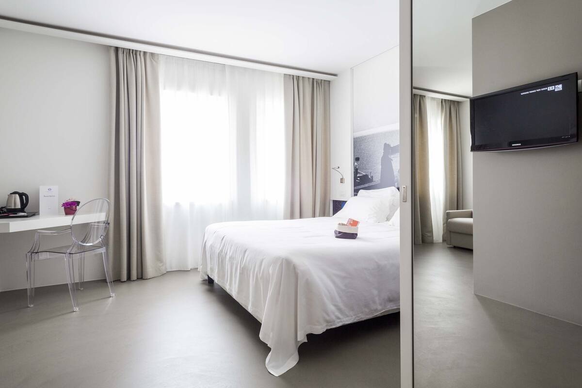 A hotel room with a bed, a desk with a chair, a television, and a window with curtains.