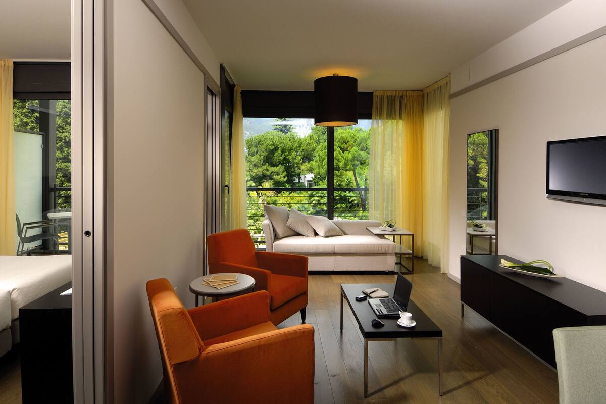 A hotel room with a bed, orange armchair, sofa, TV, and a balcony with a view of greenery.