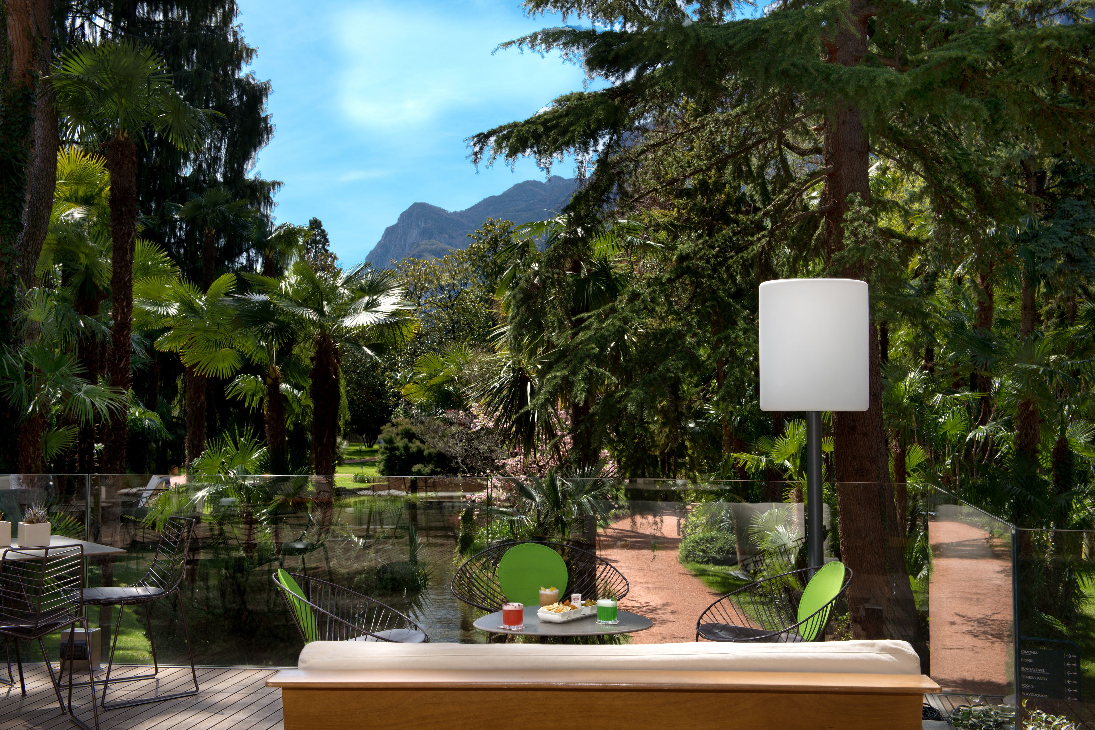 A glass-enclosed outdoor dining area with a table set for two, surrounded by lush tropical vegetation and a mountain view.