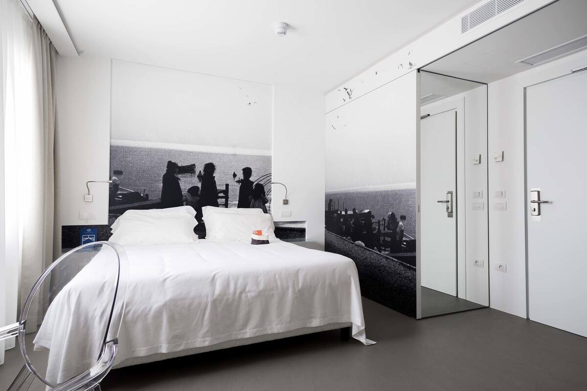A modern hotel room with a large bed, a glass-walled shower, and a view of a beach scene on the wall.