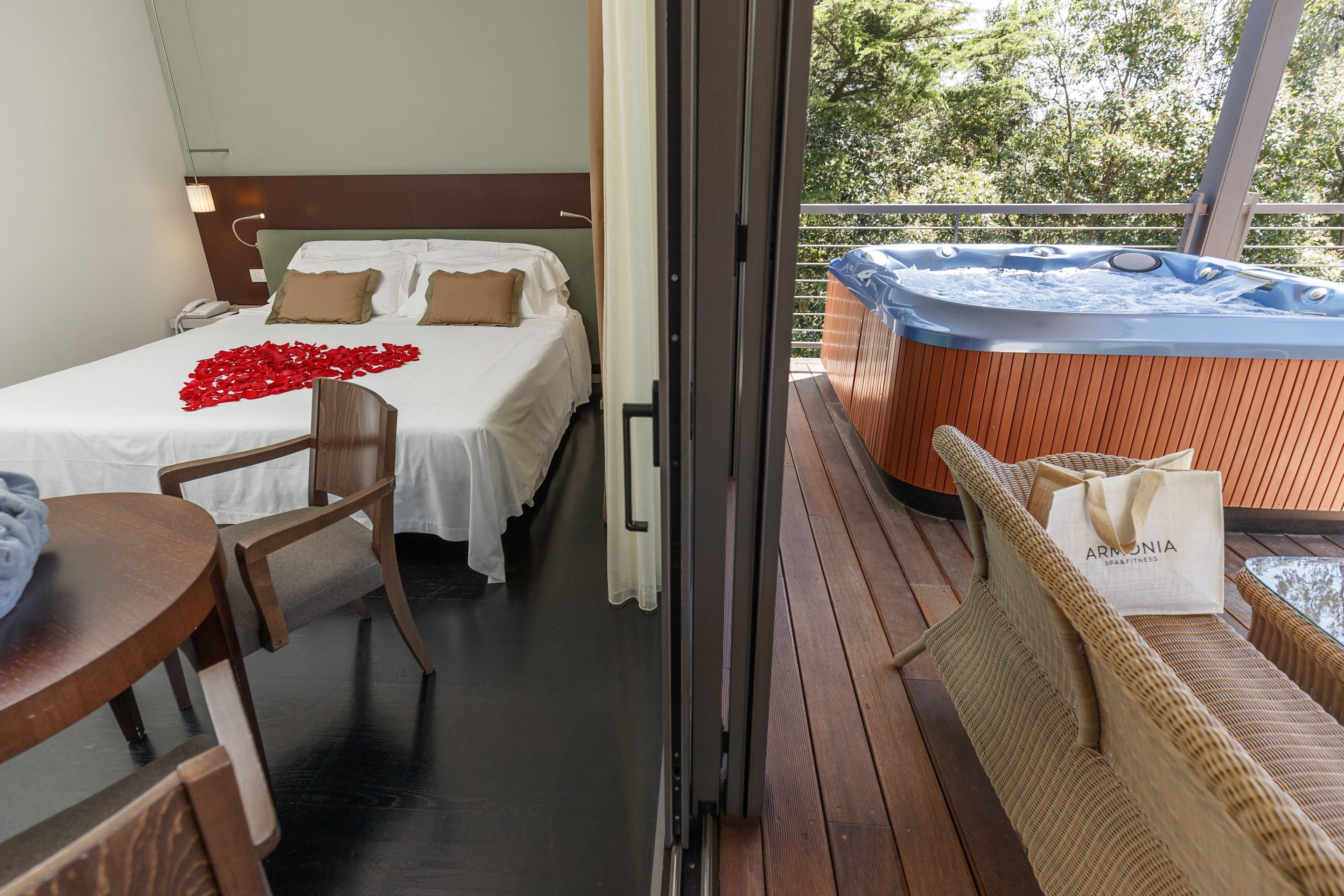 A hotel room with a bed, a chair, a table, a hot tub, and a balcony with a view of trees.