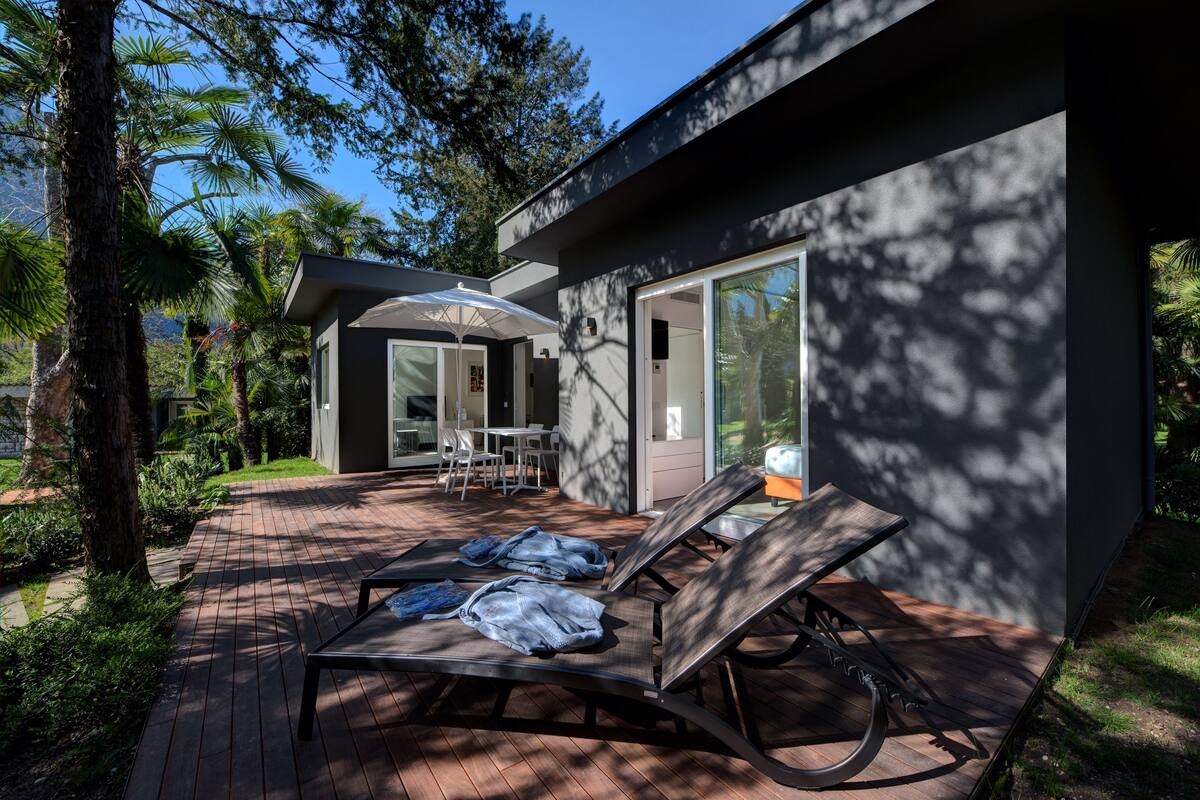 A modern outdoor patio with lounge chairs, a covered seating area, and a glass door leading to a room.