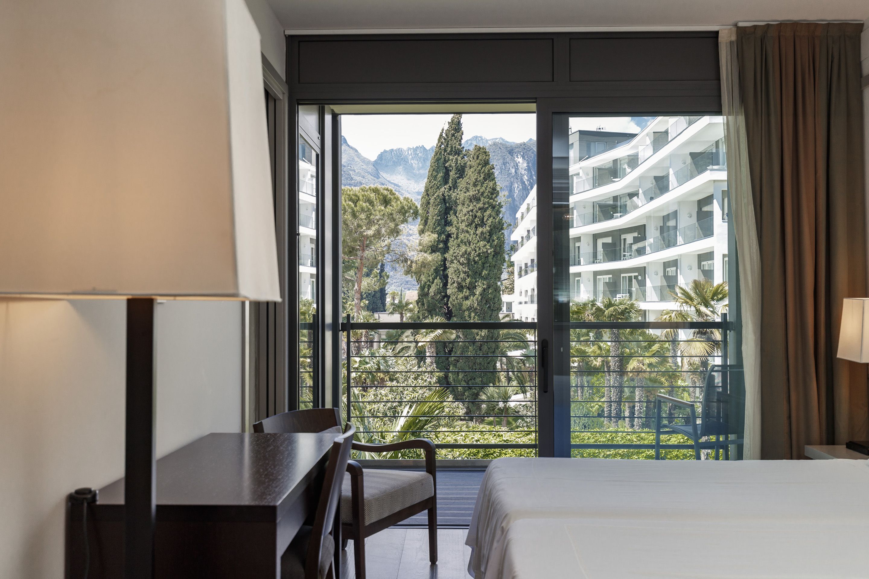 A hotel room with a bed, a desk, and a chair. The room has a large window with a view of a balcony and a mountain range.