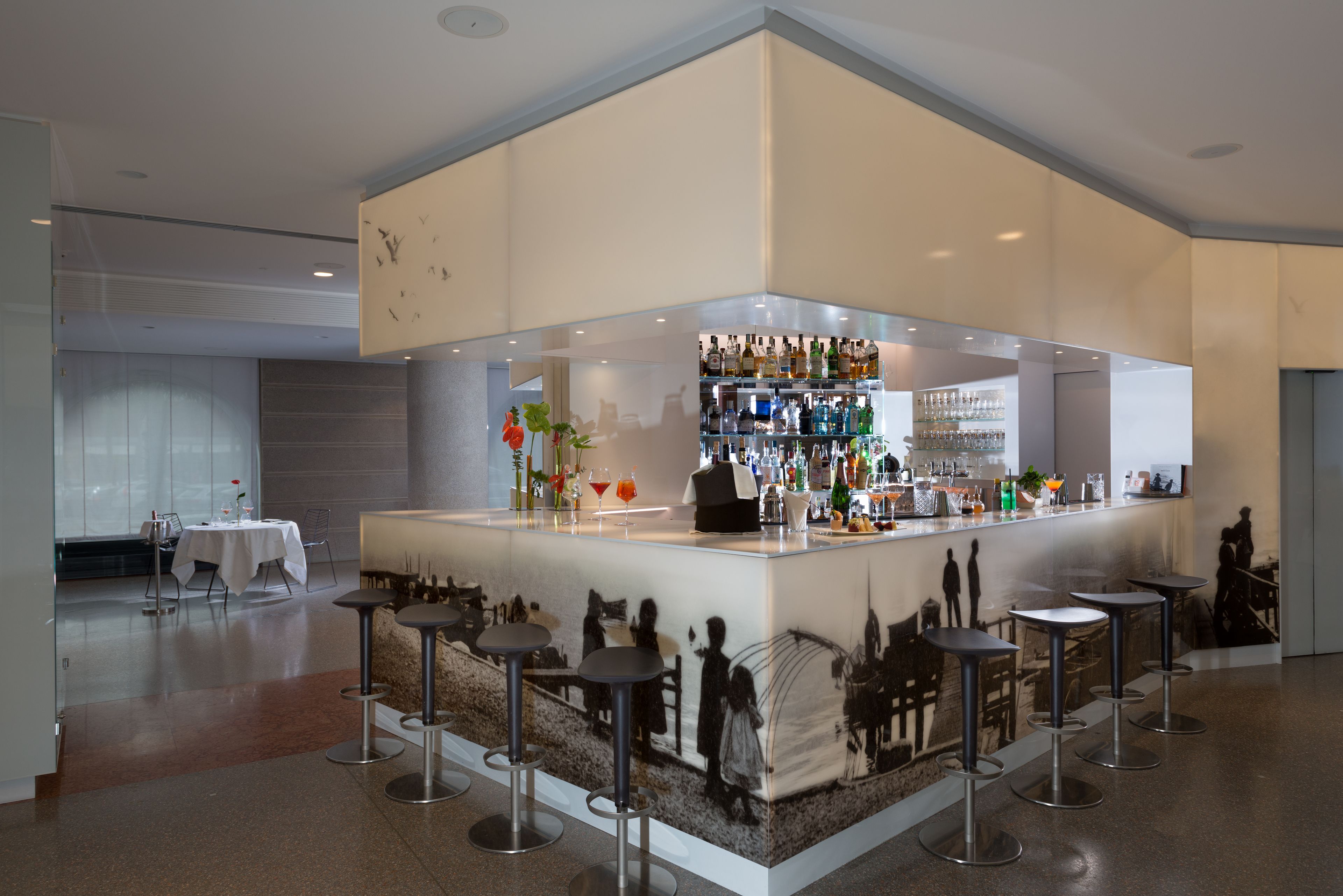 Eine Bar mit einer weißen Theke, Barhocker und einer Auswahl an Flaschen und Gläsern.