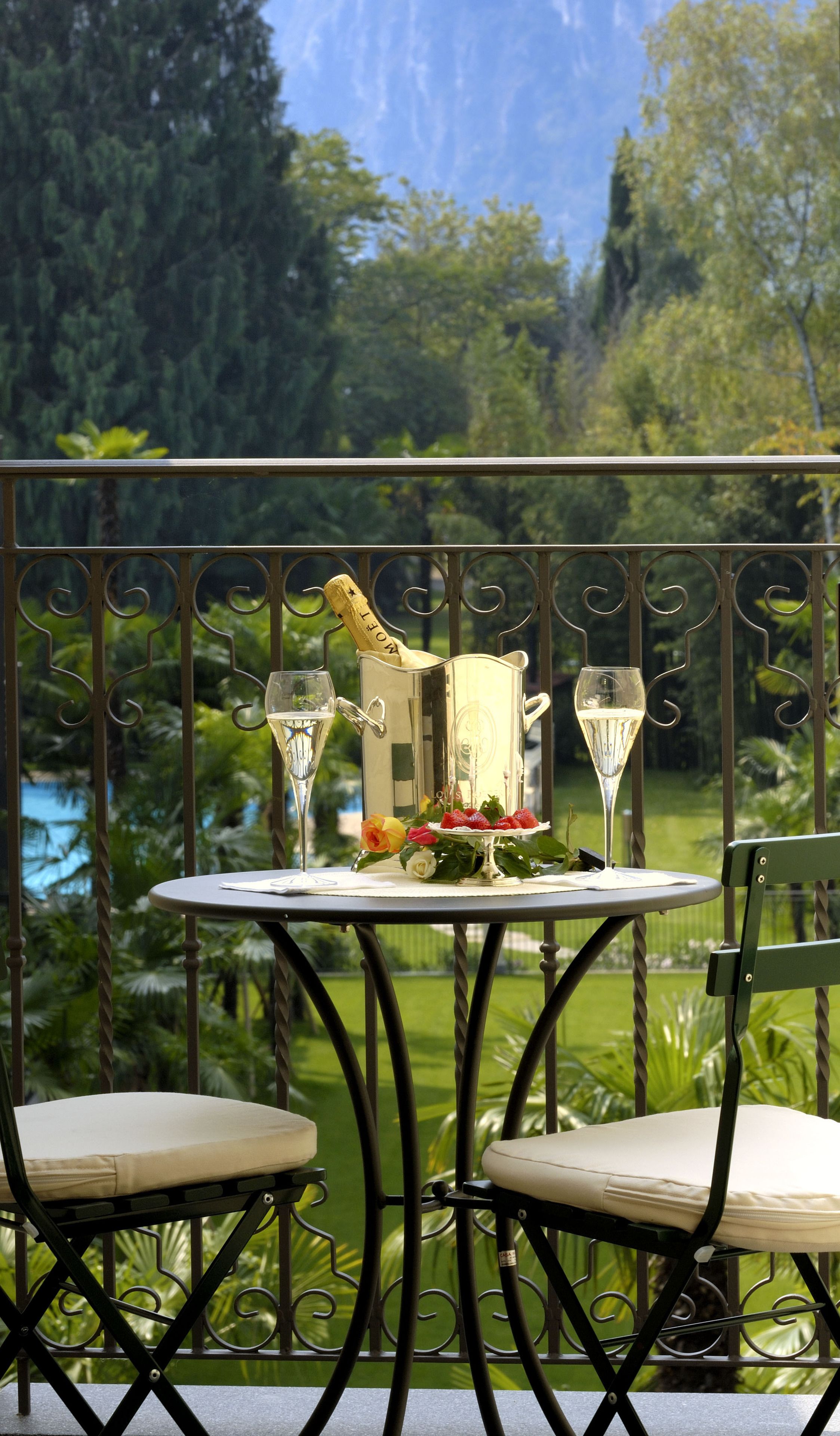 Una mesa dispuesta con una botella de champán, dos copas y un plato de fresas y rosas en un balcón con vistas a un jardín exuberante y una extensión de agua.