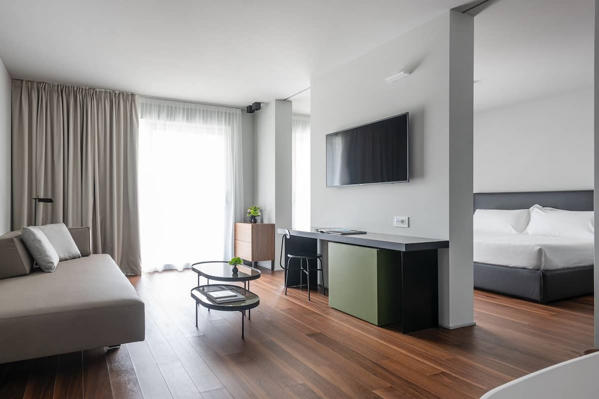 A modern hotel room with a sofa, a small round table, a desk with a chair, a bed, and a TV on the wall.
