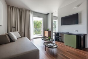 A modern living room with a sofa, a glass coffee table, a kitchen area with a green cabinet, and a large TV on the wall.