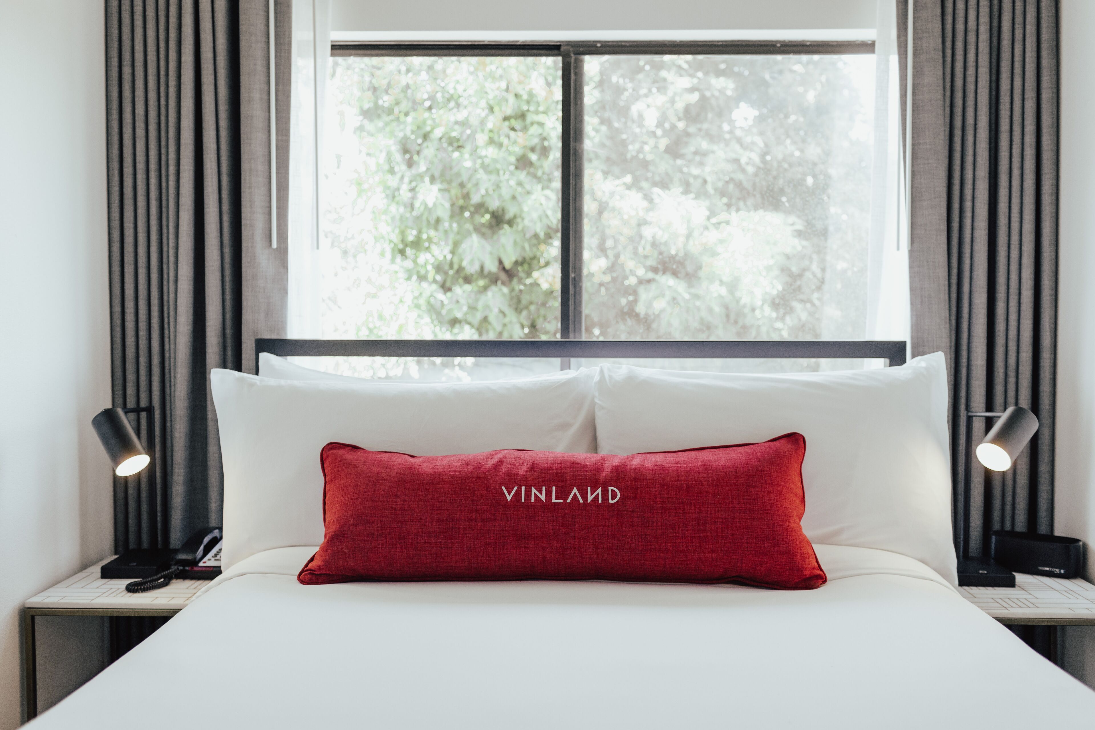 A hotel room with a bed, a red pillow with the brand name, a window with curtains, and bedside lamps.