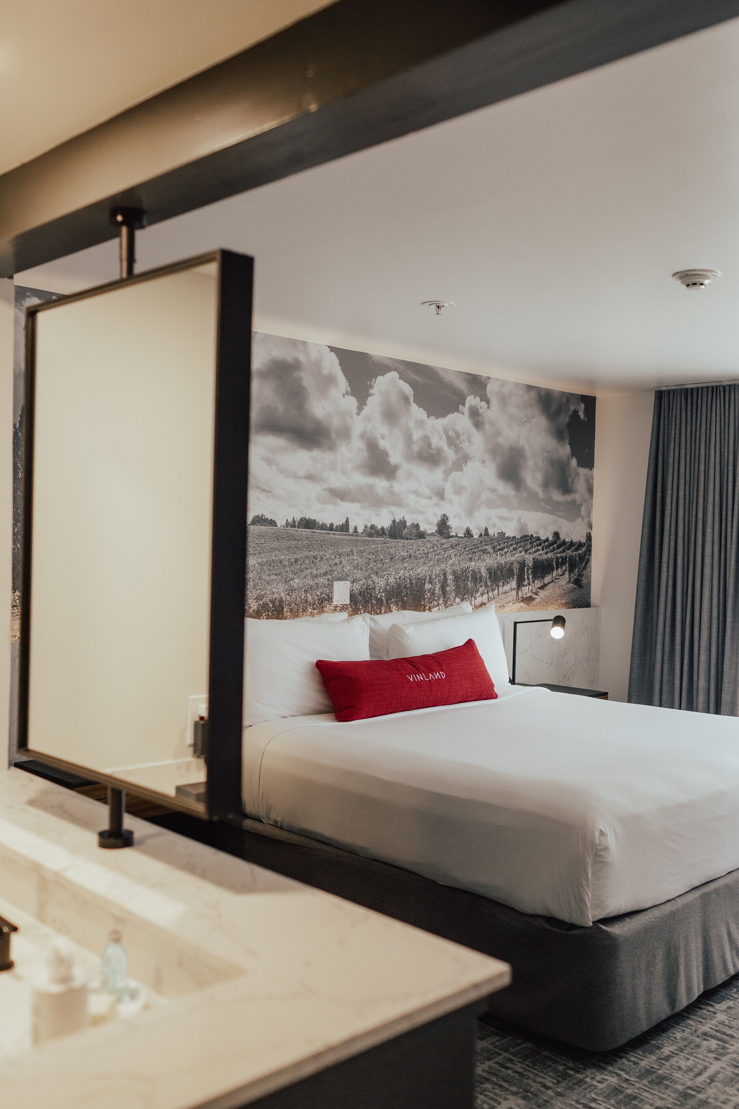 Une chambre d’hôtel avec un lit, une table de chevet, un miroir, une grande fresque murale et une fenêtre avec des rideaux.