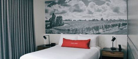 Habitación de hotel con una cama grande, una almohada roja, una mesita de noche con una lámpara y un cuadro colgado en la pared de un paisaje.