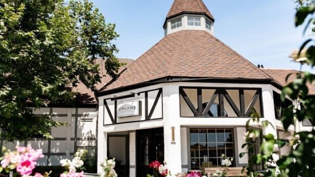 A traditional Tudor-style building with a conical roof, a sign reading "VINLAND," and a garden with various flowers in the foreground.