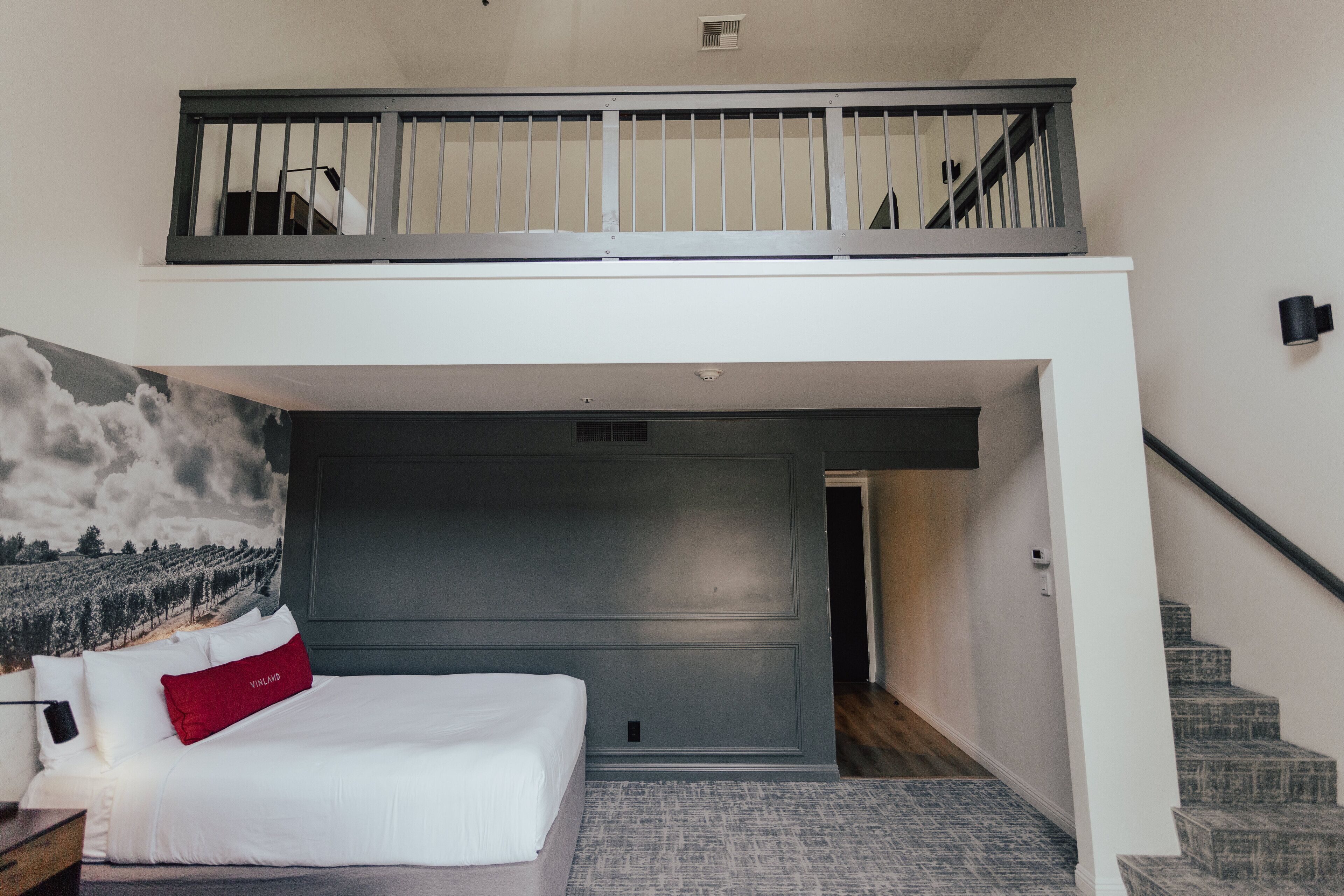 Un dormitorio con una cama grande, un almohadón rojo, un balcón con barandilla y una escalera que sube a un nivel superior.