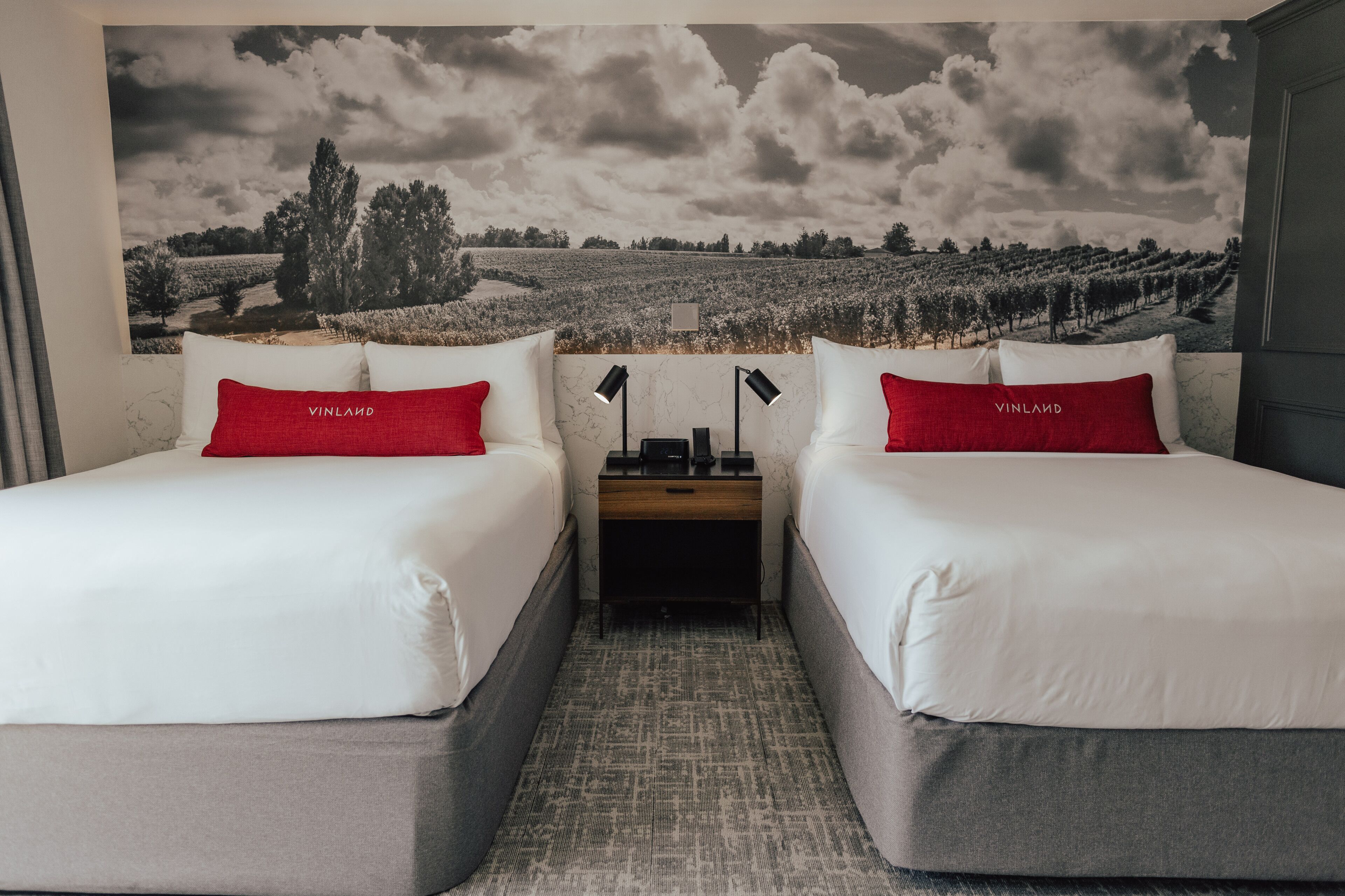 Habitación de hotel con dos camas, cada una con un almohadón rojo y una colcha blanca. Entre las camas hay una mesita de noche con una lámpara y una pequeña mesa. Un mural grande muestra un paisaje de viñedo.