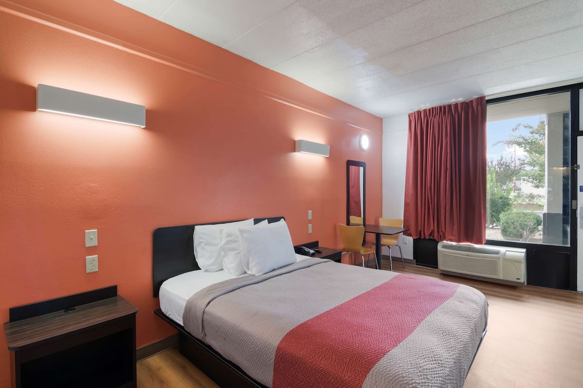 A hotel room with a bed, bedside table, a desk with a chair, a window with curtains, and a view of the outdoors.