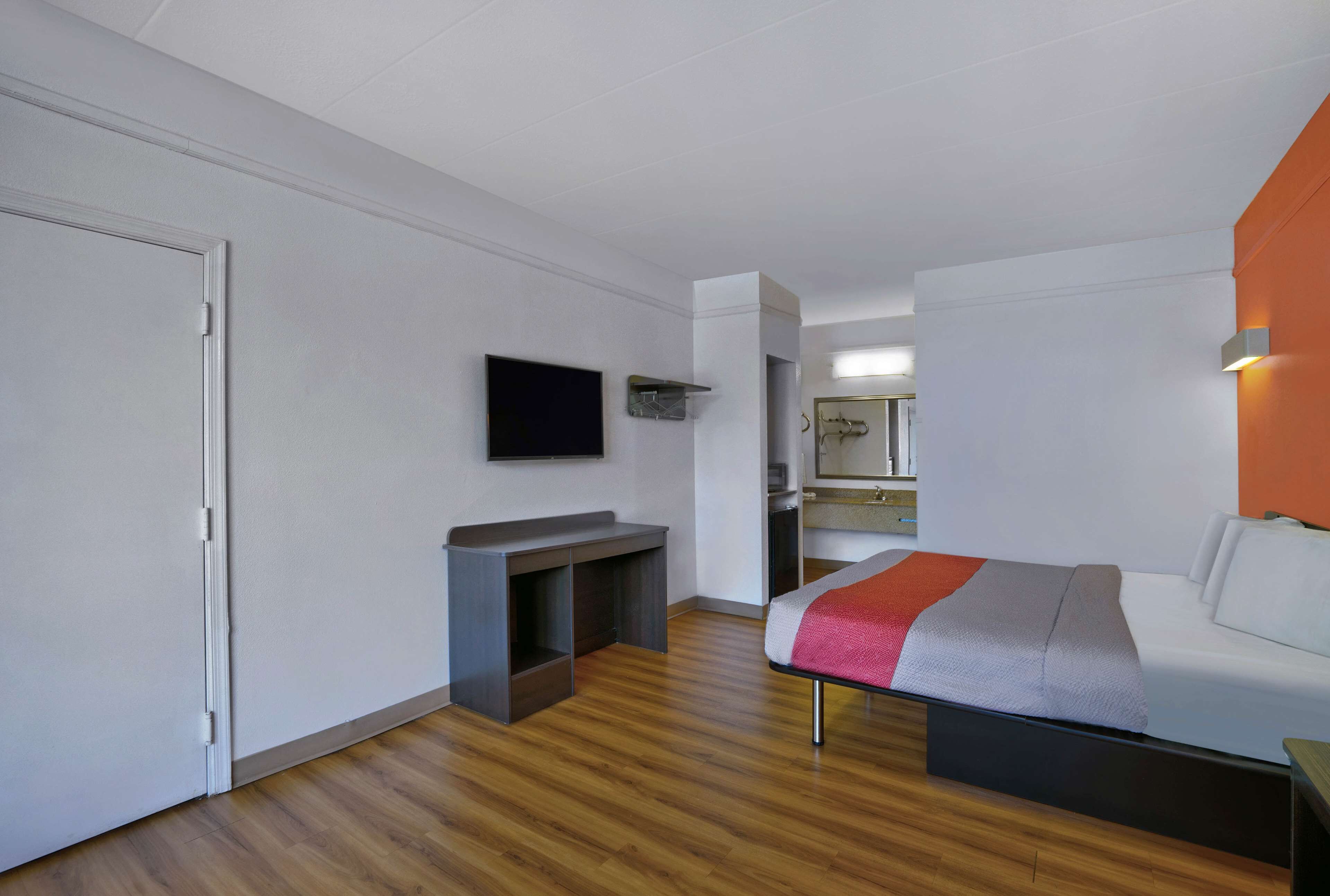 Una habitación de hotel moderna con piso de madera, una cama con manta gris y roja, un televisor de pantalla plana montado en la pared y un pequeño escritorio con espejo.