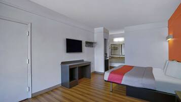Una habitación de hotel moderna con piso de madera, una cama con manta gris y roja, un televisor de pantalla plana montado en la pared y un pequeño escritorio con espejo.