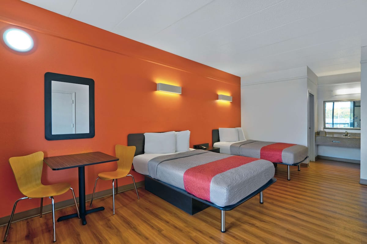 A hotel room with two beds, a dining table, chairs, a mirror, and a bathroom visible through an open door.