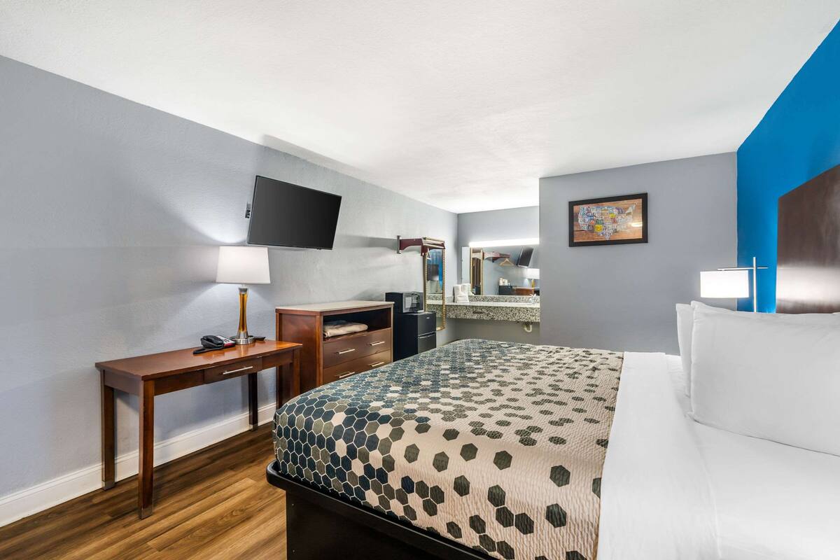A hotel room with a bed, a desk, a chair, a TV, a mirror, and a bathroom area.