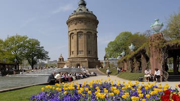 Een historische toren met een koepel, omringd door een bloementuin met gele en paarse bloemen, en een geplaveid pad.