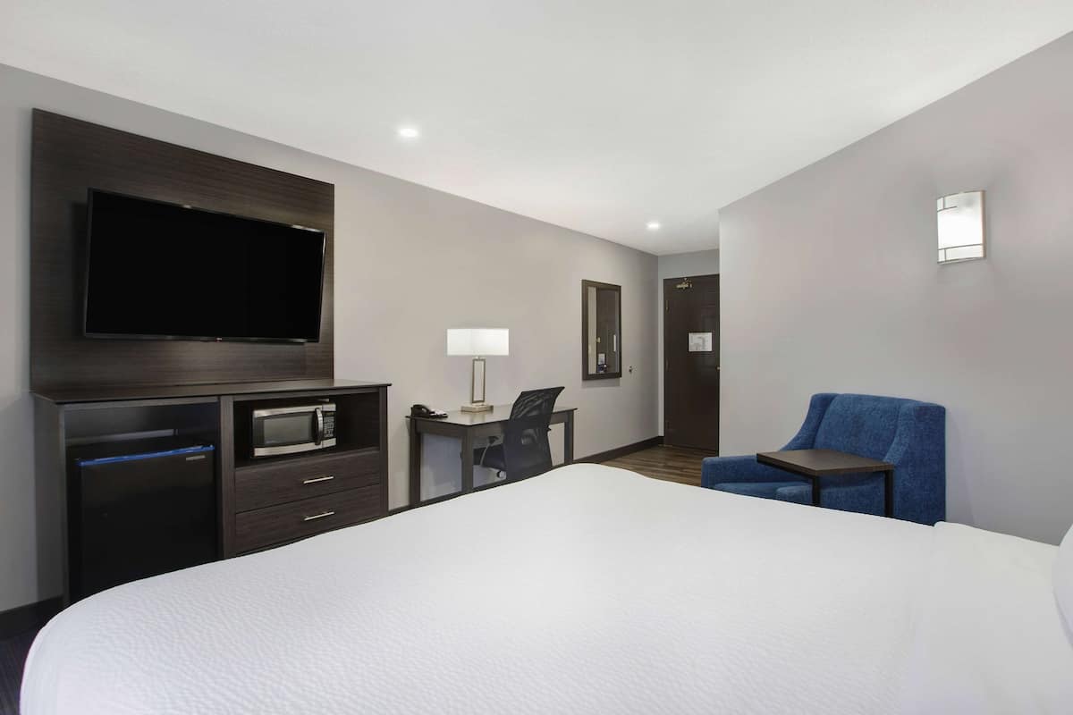 A hotel room with a bed, a flat-screen TV, a desk with a lamp, a microwave, and a blue chair.