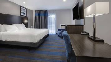 A hotel room with a bed, a desk, a chair, a flat-screen TV, a lamp, and a window with curtains.