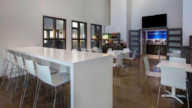 A modern dining area with a long white table and multiple white chairs. There is a counter with a coffee machine and a sign that says "Breakfast is on."