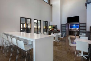 Un comedor moderno con una mesa blanca larga y múltiples sillas blancas. Hay una barra con una máquina de café y un cartel que dice "Desayuno incluido".