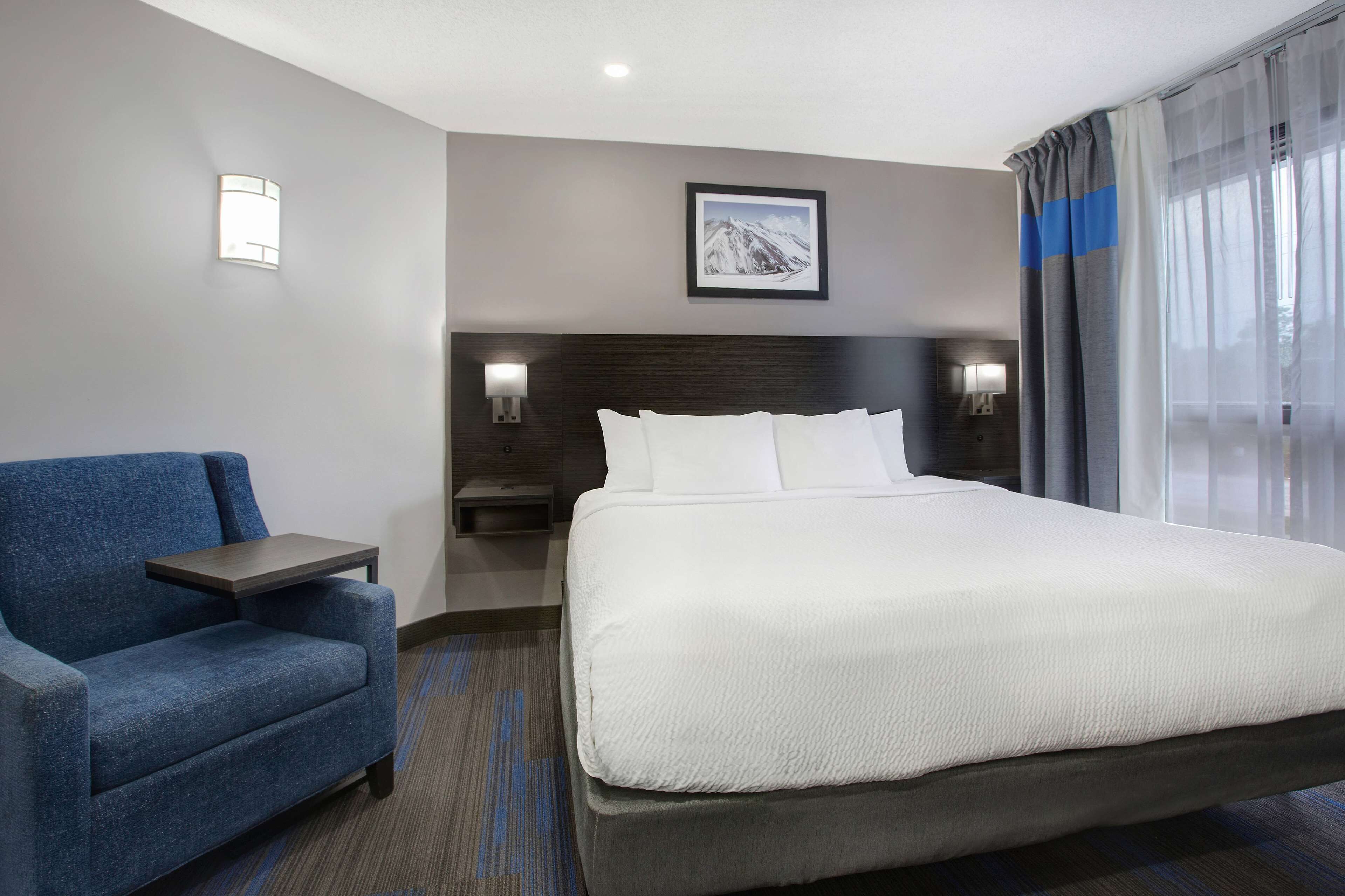 Habitación de hotel con una cama grande, un sillón azul, una mesita de noche y un cuadro de una montaña en la pared.