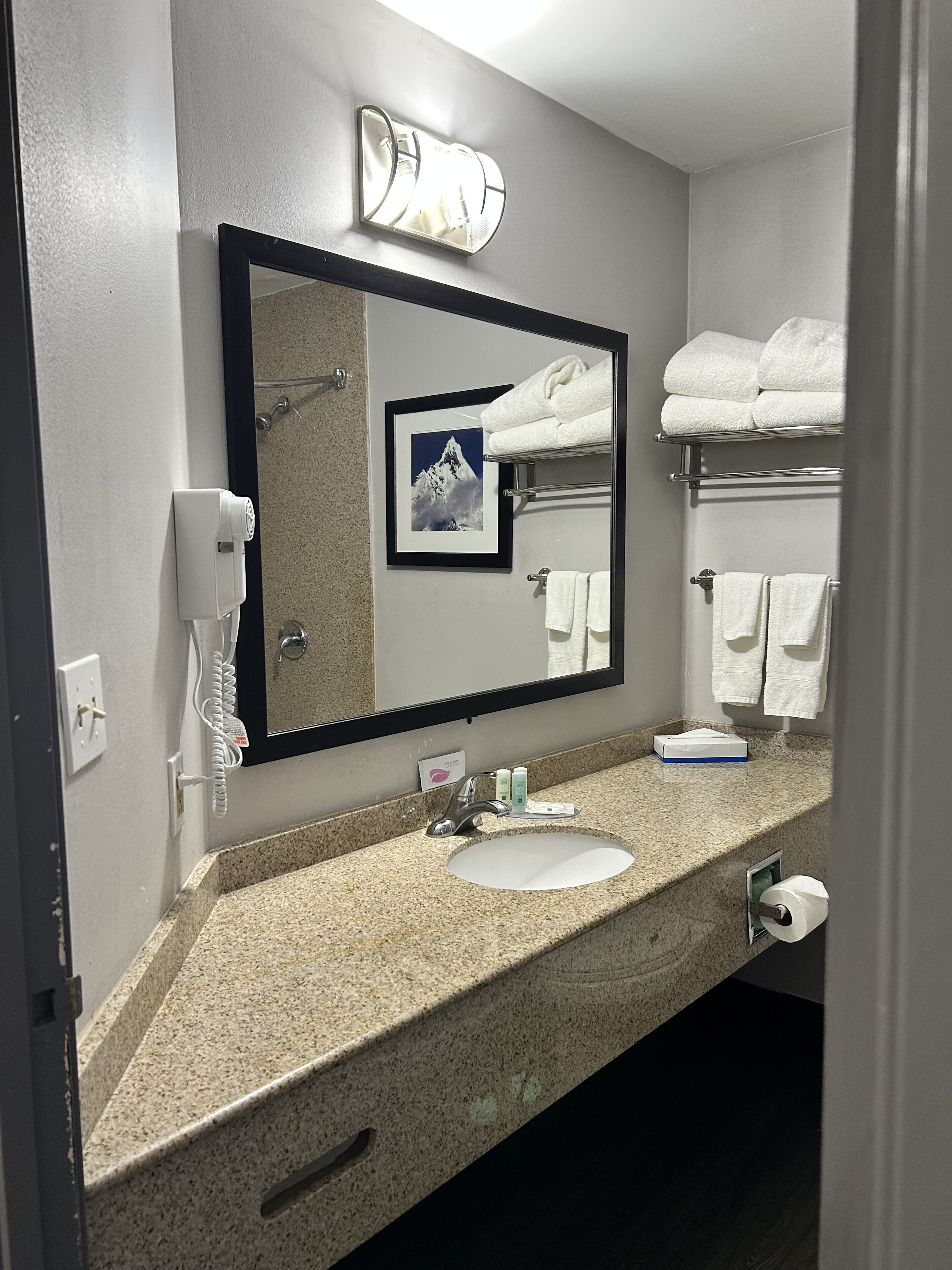 A bathroom with a granite countertop, a sink, a mirror, a towel rack with folded towels, a framed picture, a hairdryer, and a phone.