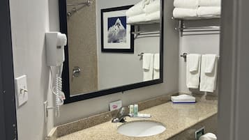 A bathroom with a granite countertop, a sink, a mirror, a towel rack with folded towels, a framed picture, a hairdryer, and a phone.