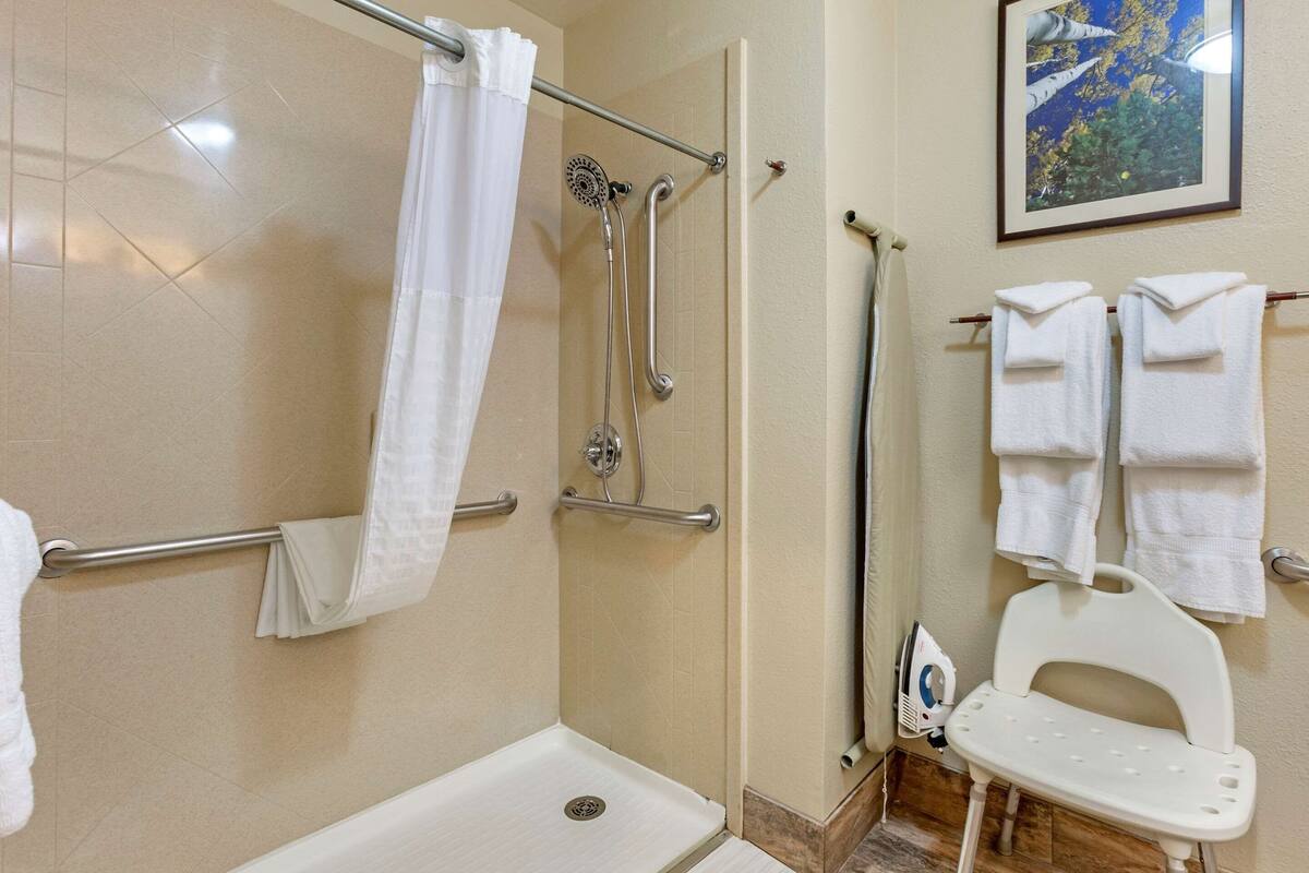 A bathroom with a shower, grab bars, a white bathtub, a white toilet, and a white bench.