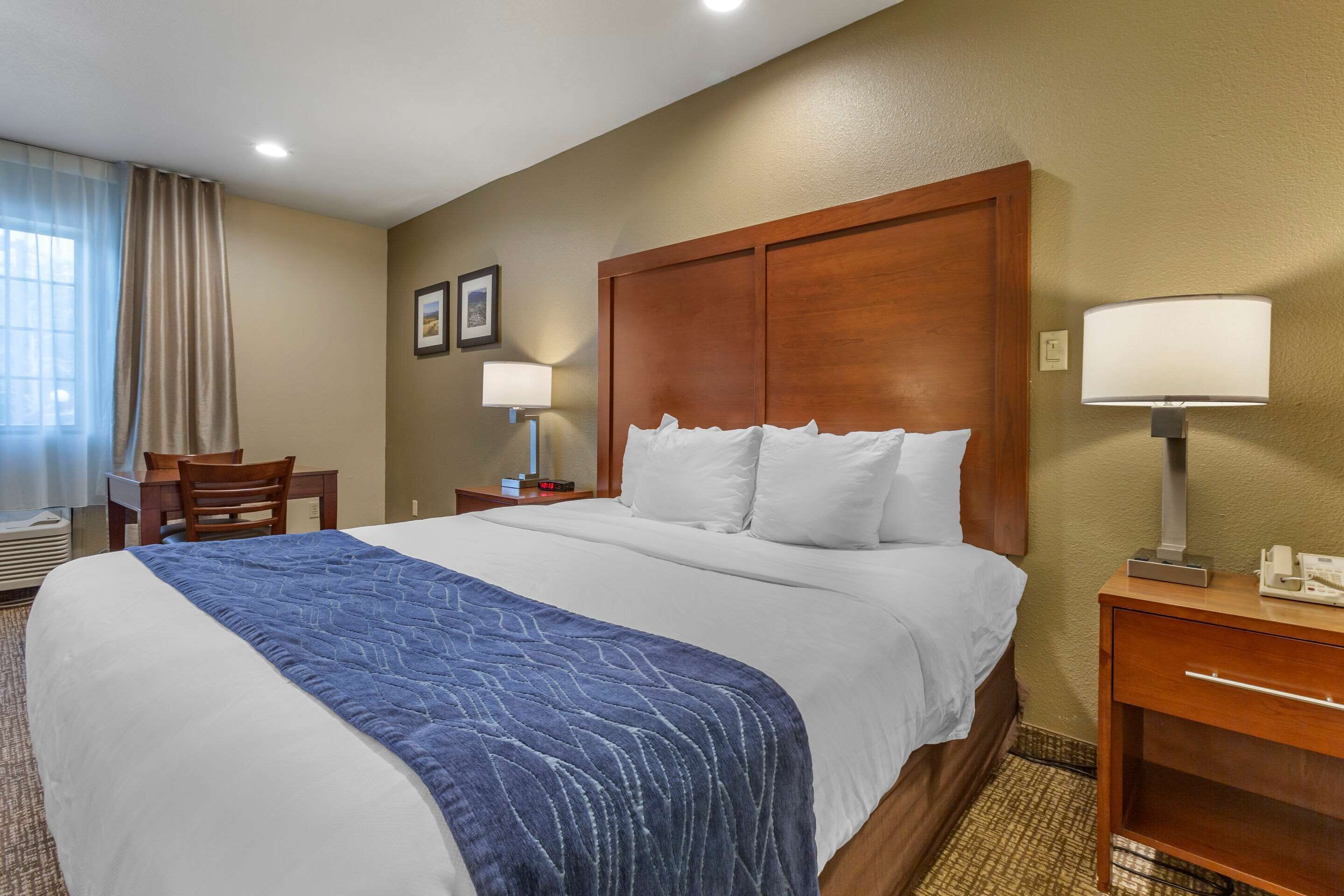 A hotel room with a large bed, a wooden headboard, a desk with a chair, a nightstand with a lamp, and a window with curtains.