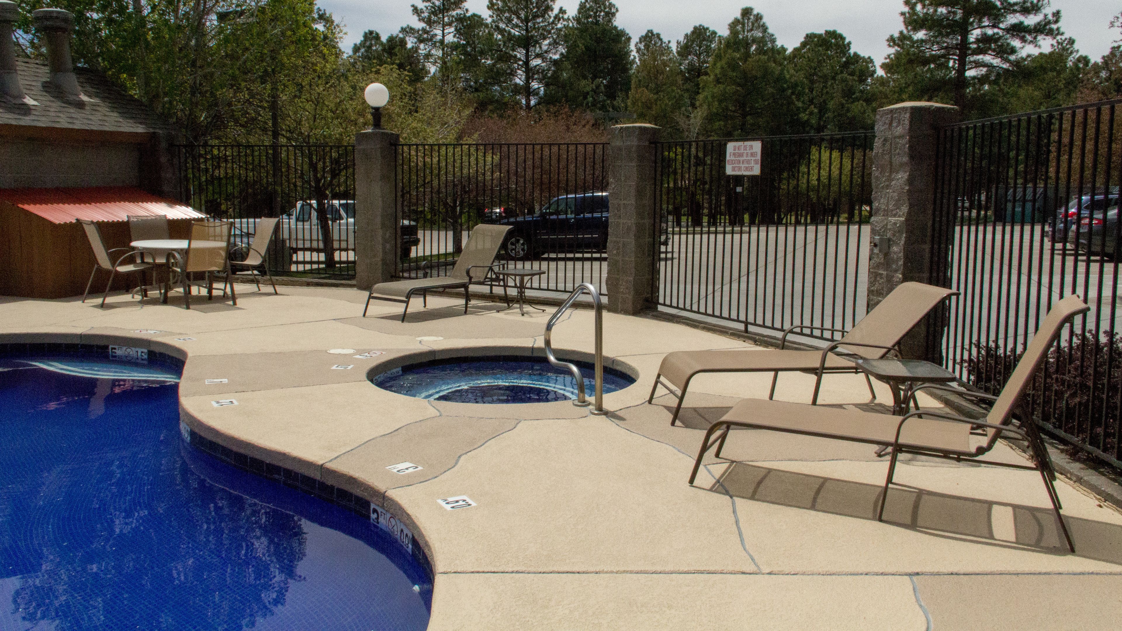 A pool area with lounge chairs, a hot tub, and a fenced parking area.