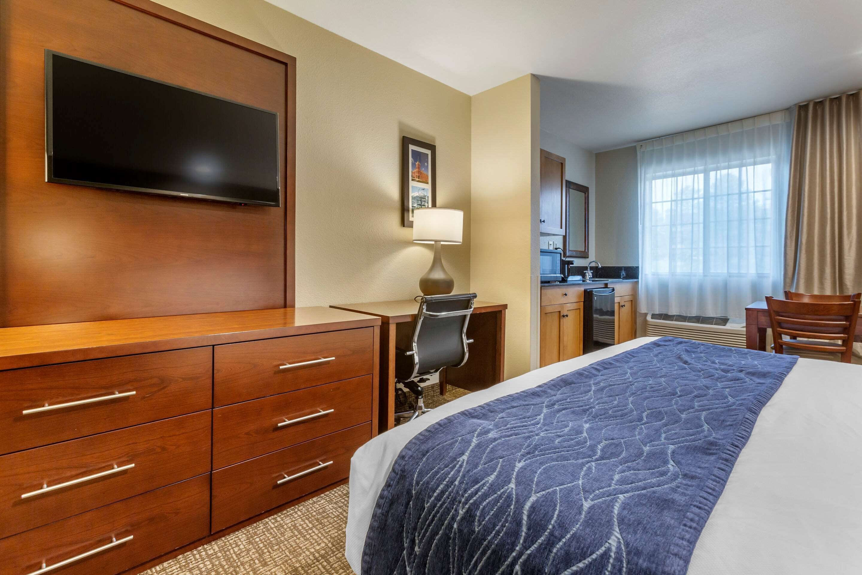 Een hotelkamer met een groot bed, een bureau met een stoel, een flatscreen televisie, een lamp en uitzicht op de keuken en badkamer door het raam.